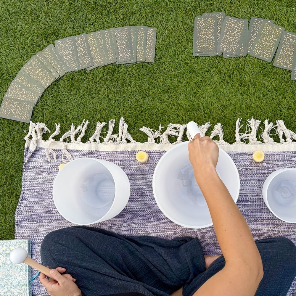 Overhead view of crystal singing bowls on a woven blanket, a hand playing one with a mallet, oracle cards fanned out on the grass