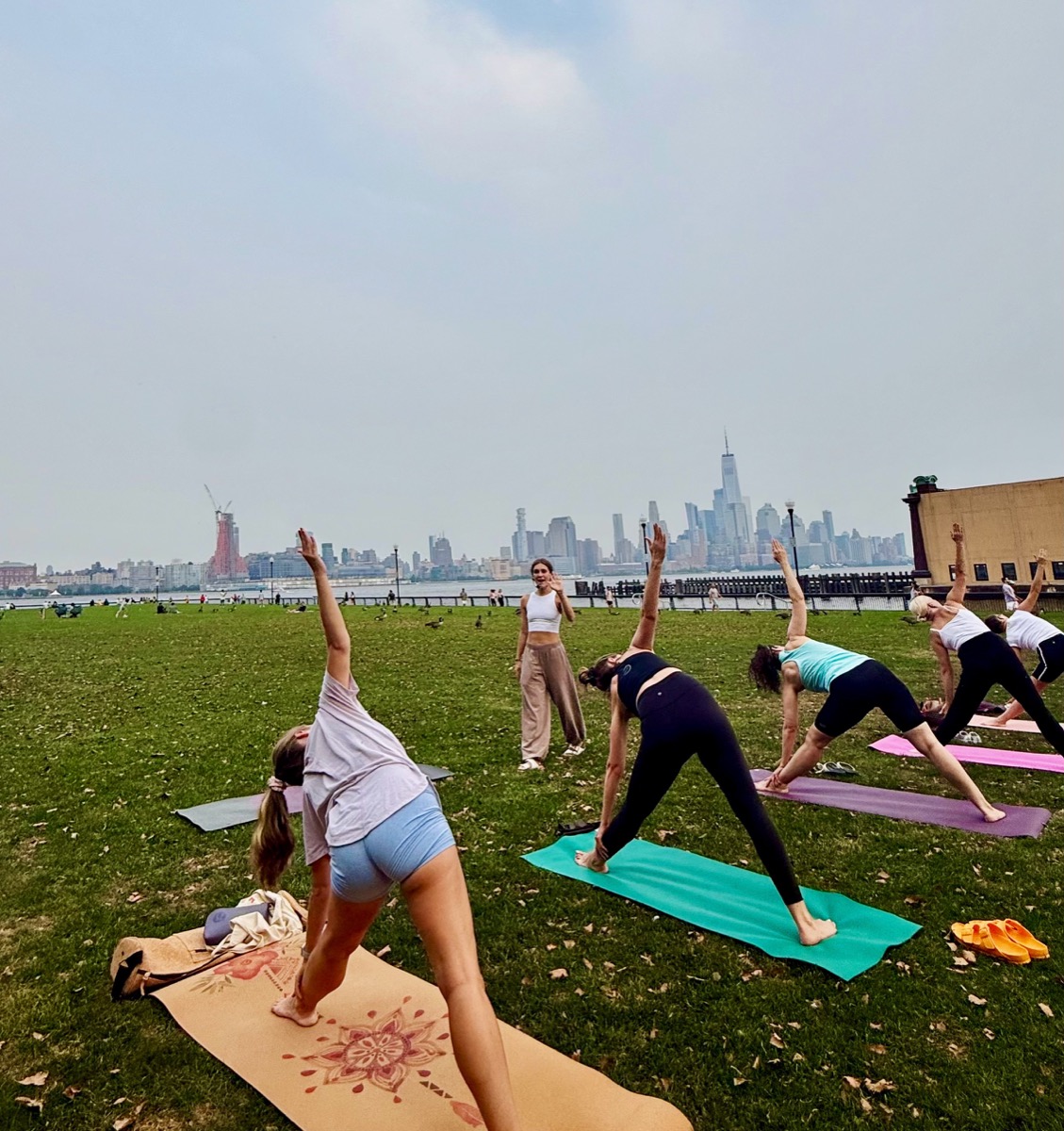 A group in triangle pose on the lawn at the Hoboken waterfront, with the Manhattan skyline across the river