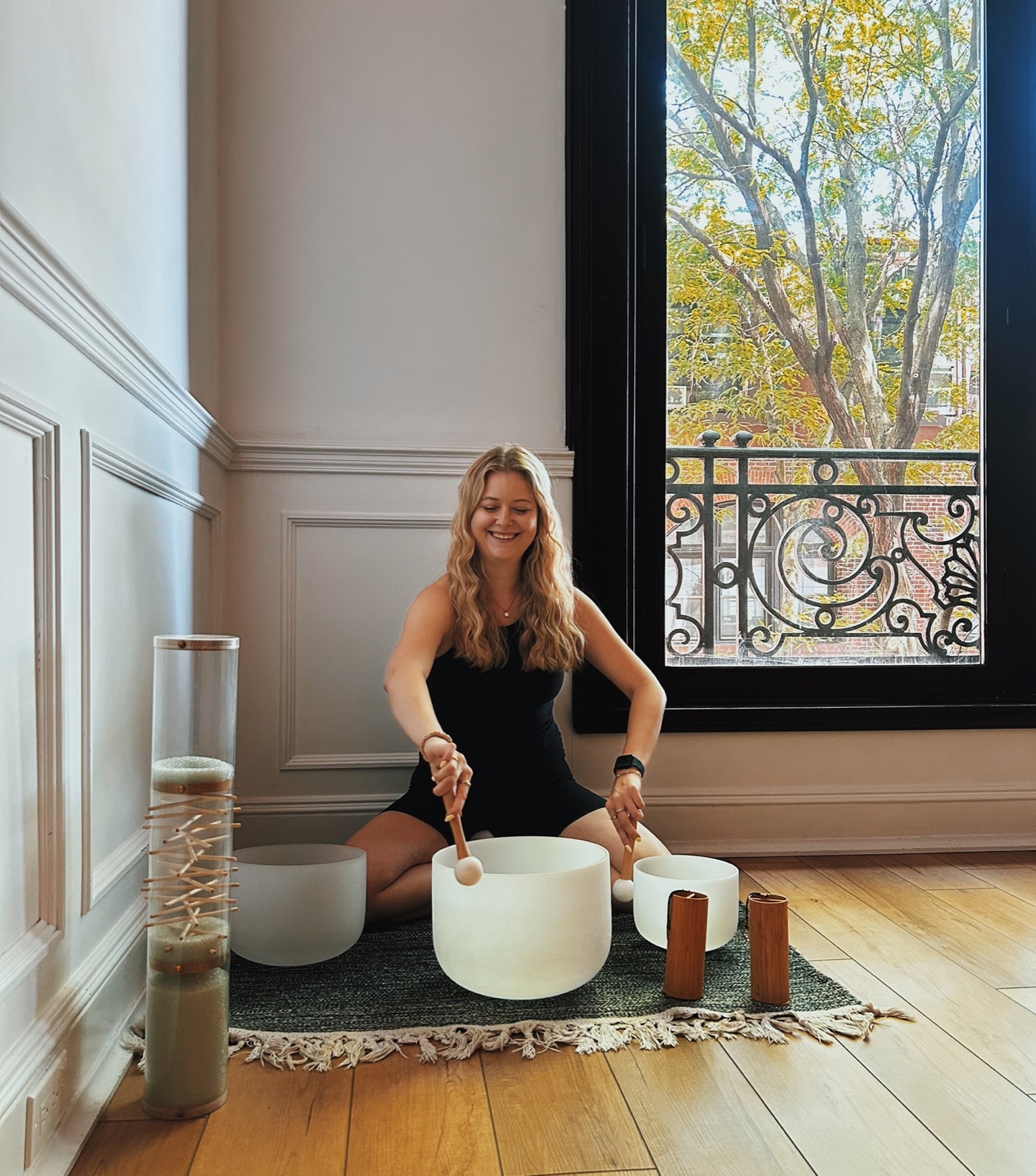 A practitioner seated on a rug playing three crystal singing bowls beside a tall window with an iron balcony and autumn trees